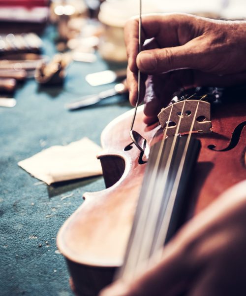 Senior craftsman checking the violin in his workshop Portrait of senior entrepreneur working in his instrument repair shop, taking care of violins and other music instruments. Experienced senior is using his skills to repair instruments